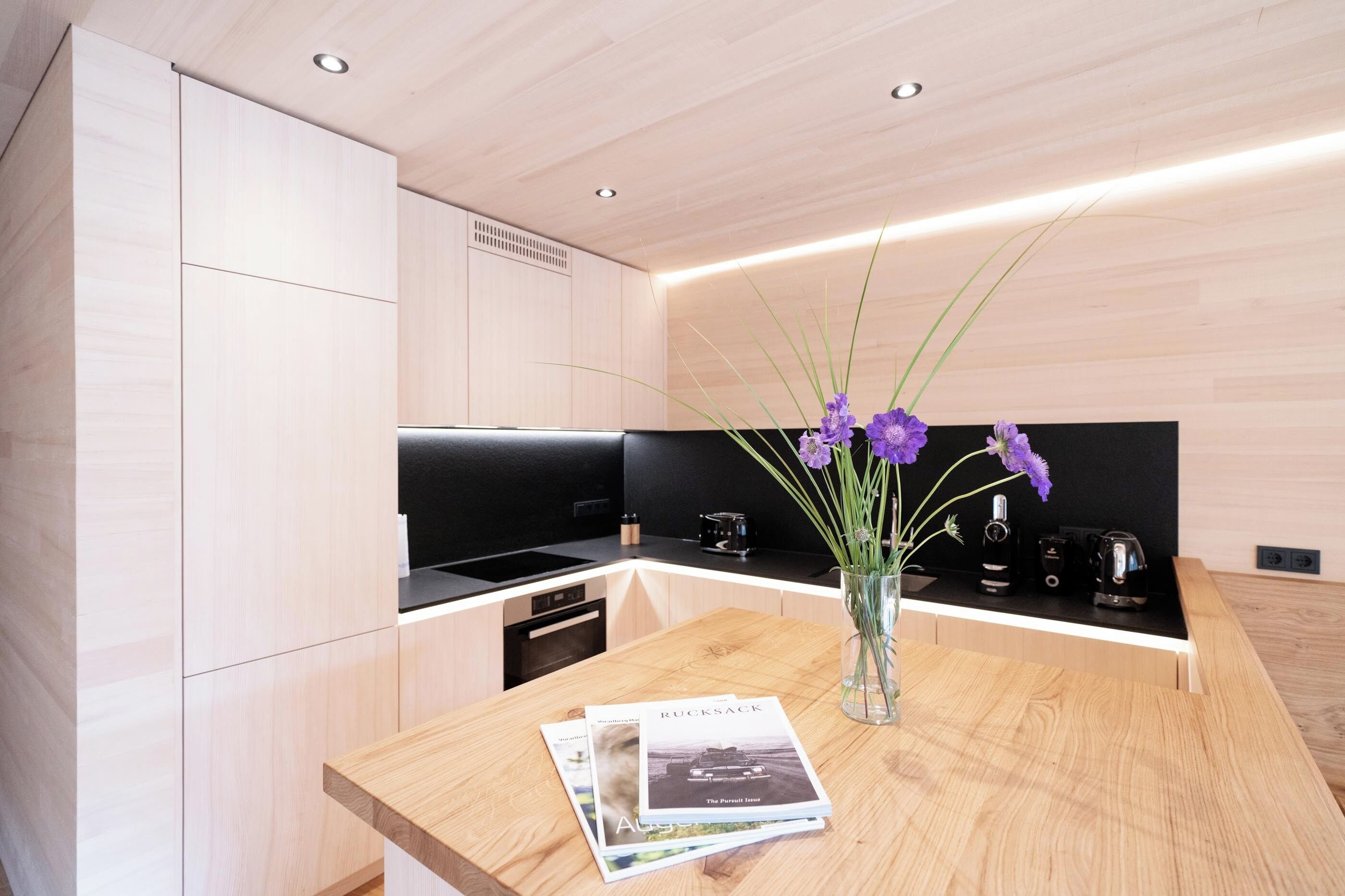 A modern kitchen with wooden cabinetry, a black countertop, and a vase with purple flowers on a wooden table.