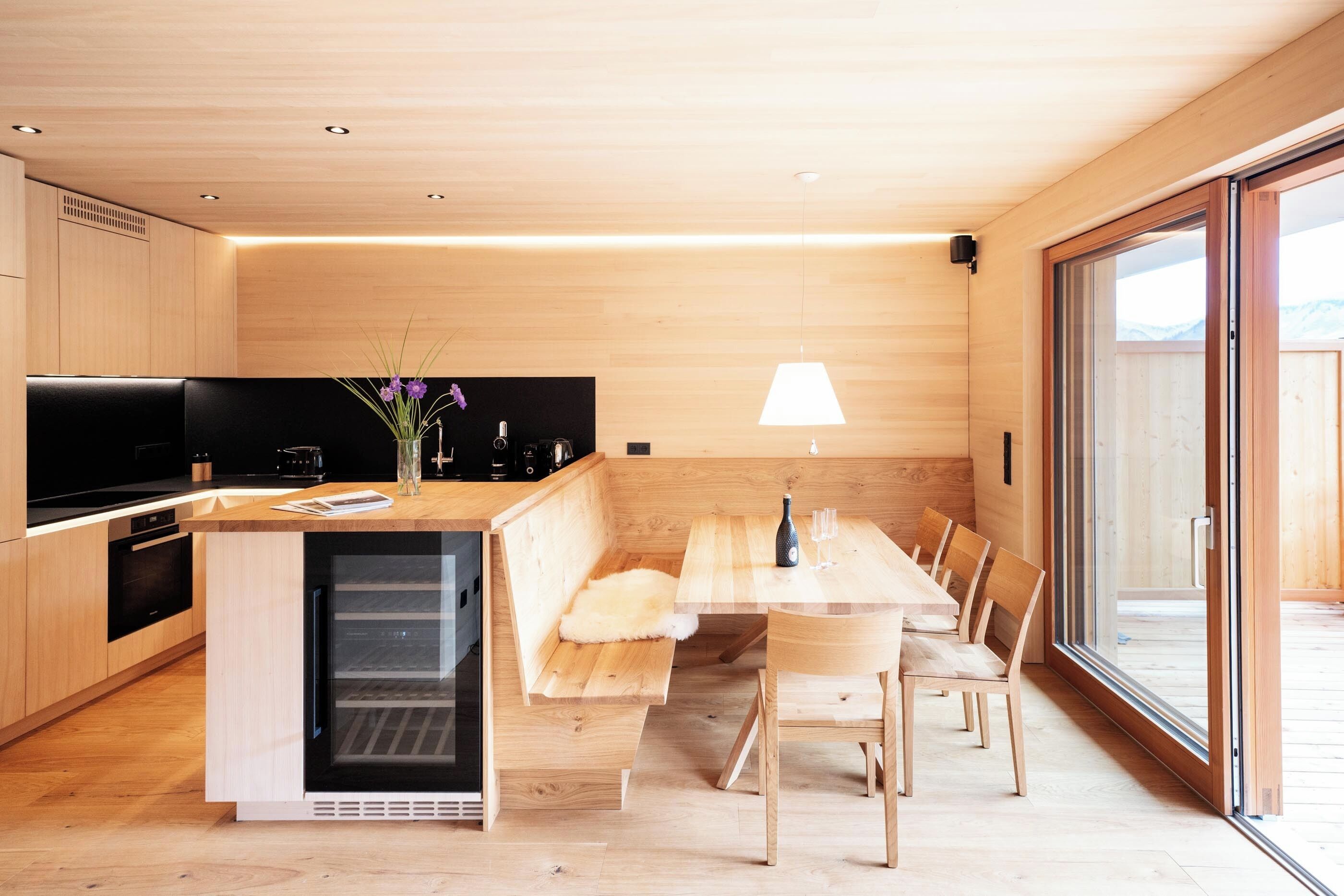 A modern kitchen with a built-in oven, a dining area with a bench, and a large window.