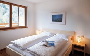 Una cama doble con sábanas blancas, un cuadro enmarcado en la pared y una ventana con vistas a árboles.