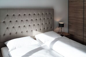 A bed with a tufted headboard, white bedding, and a bedside table with a lamp. - Landhaus Damüls (Damüls)