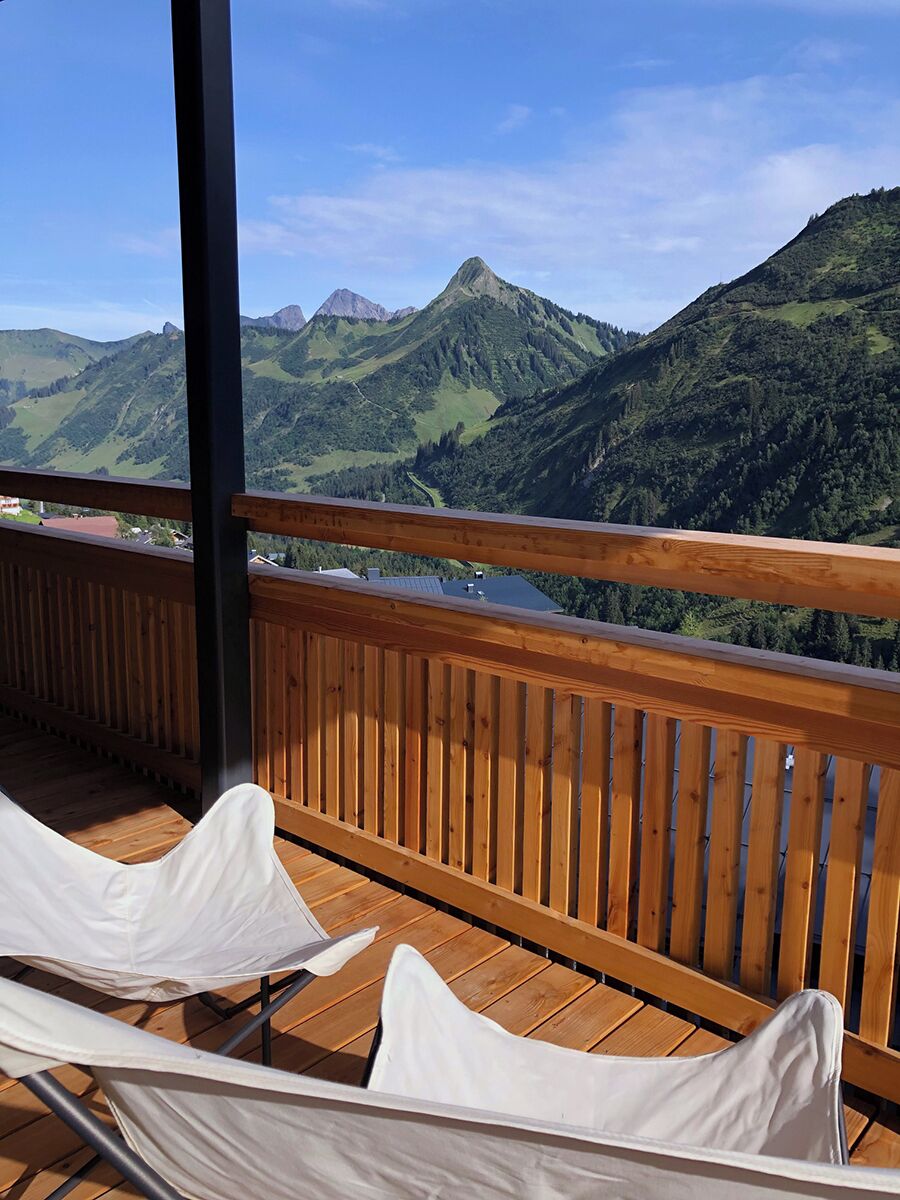 A balcony with wooden railings and white cushioned chairs, overlooking a mountainous landscape.