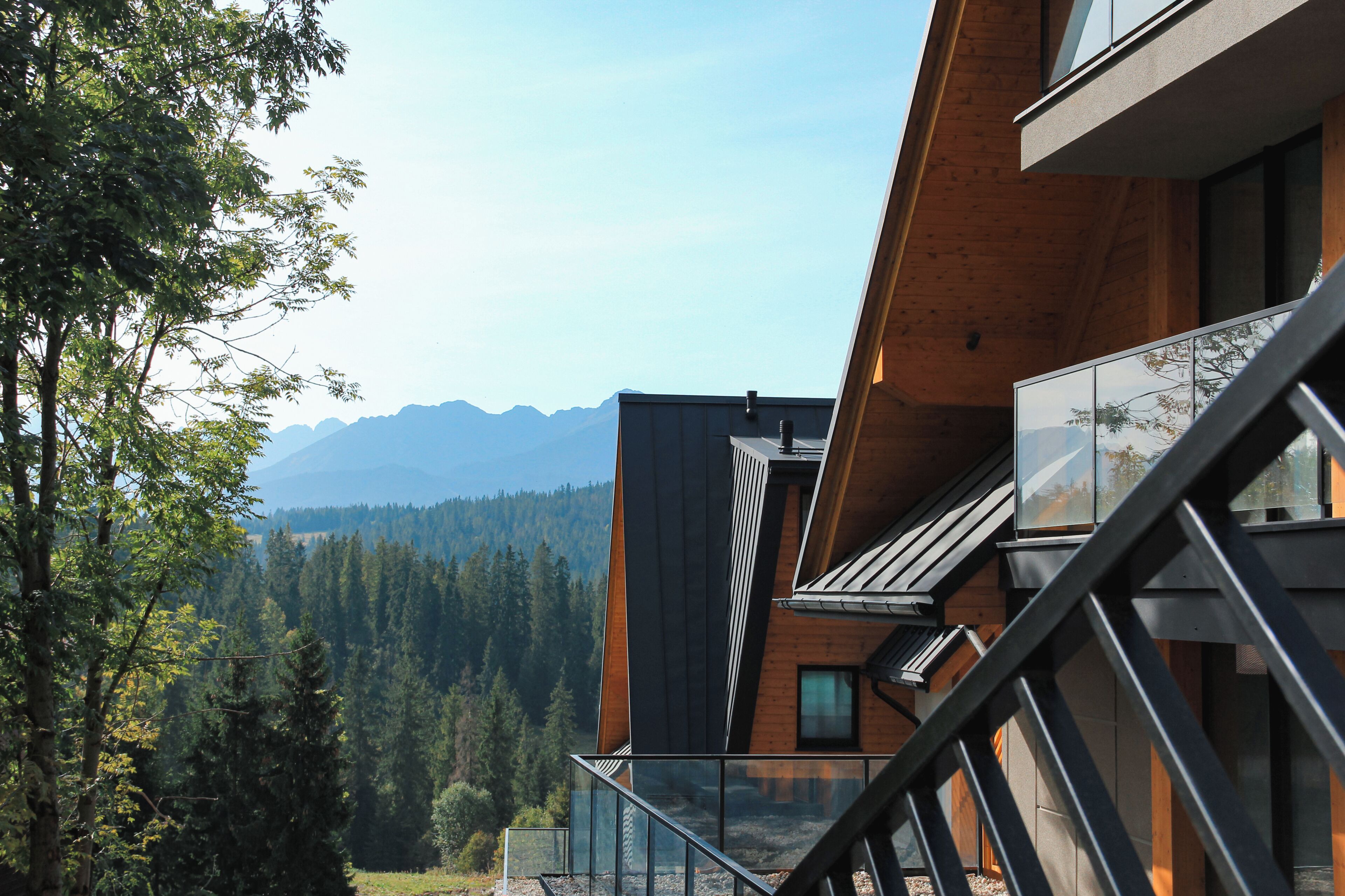 A modern building with a glass balcony overlooking a forested valley and mountains.