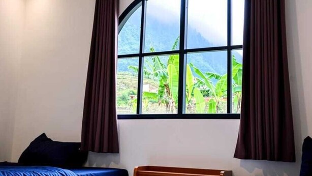 Una habitación con dos camas, un ventanal con cortinas, una lámpara colgante y vistas al exterior con vegetación.