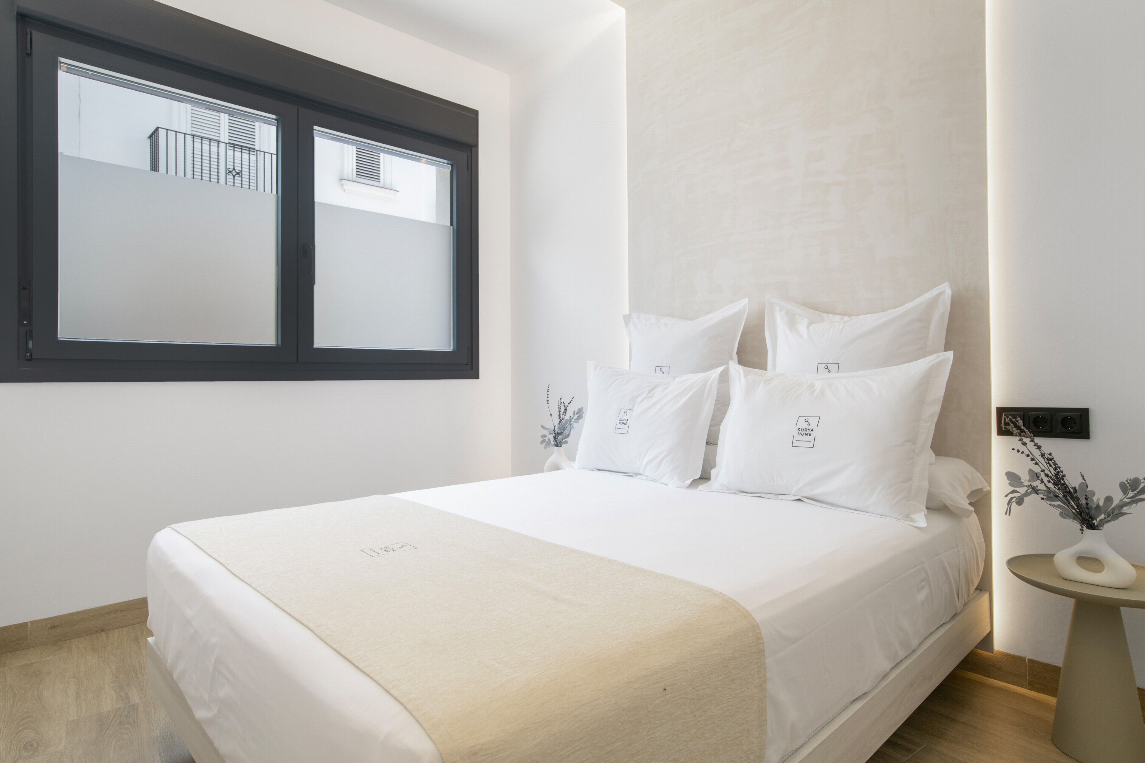 A modern bedroom with a bed, a window, a bedside table, and a wall-mounted light fixture.
