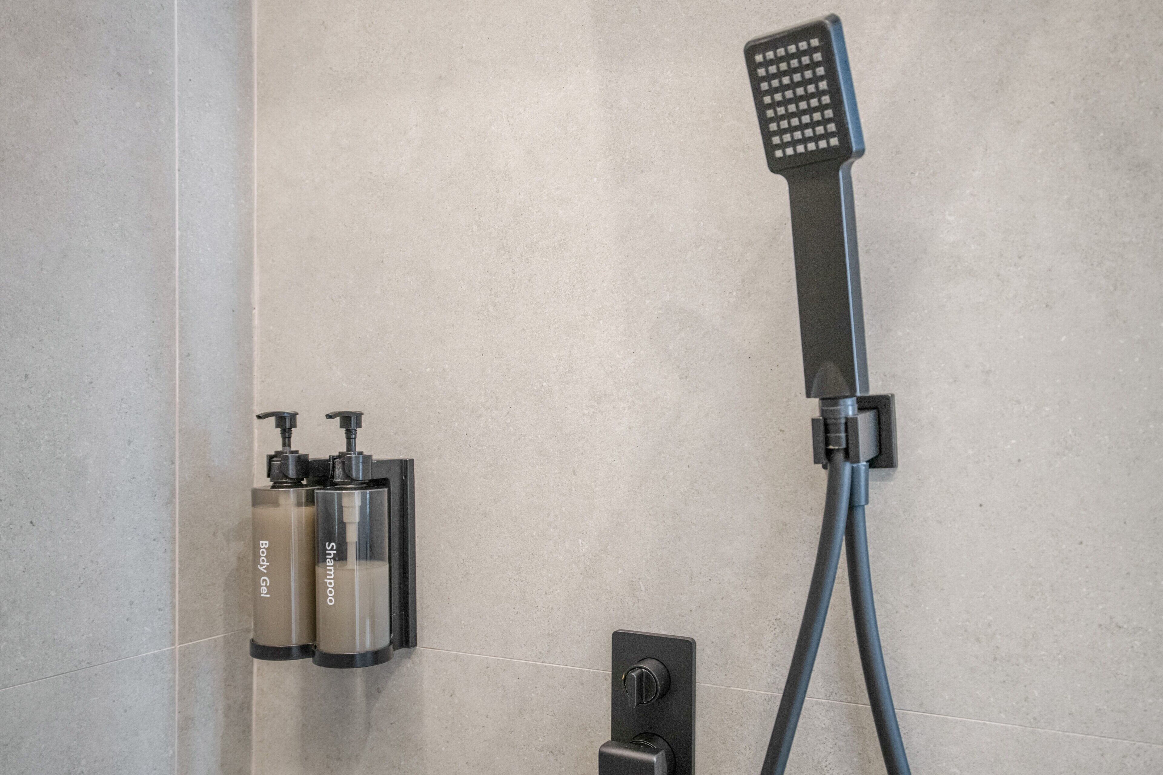 A shower area with a handheld showerhead, a wall-mounted control, and two bottles of shampoo and body gel.