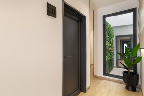 Ein Flur mit einer dunklen Tür, einem Nummernschild, einem Fenster mit Blick auf Grünflächen und einem Blumentopf.