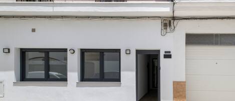 A two-story building with a white facade, two windows with black frames, a garage door, and a balcony with metal railings.