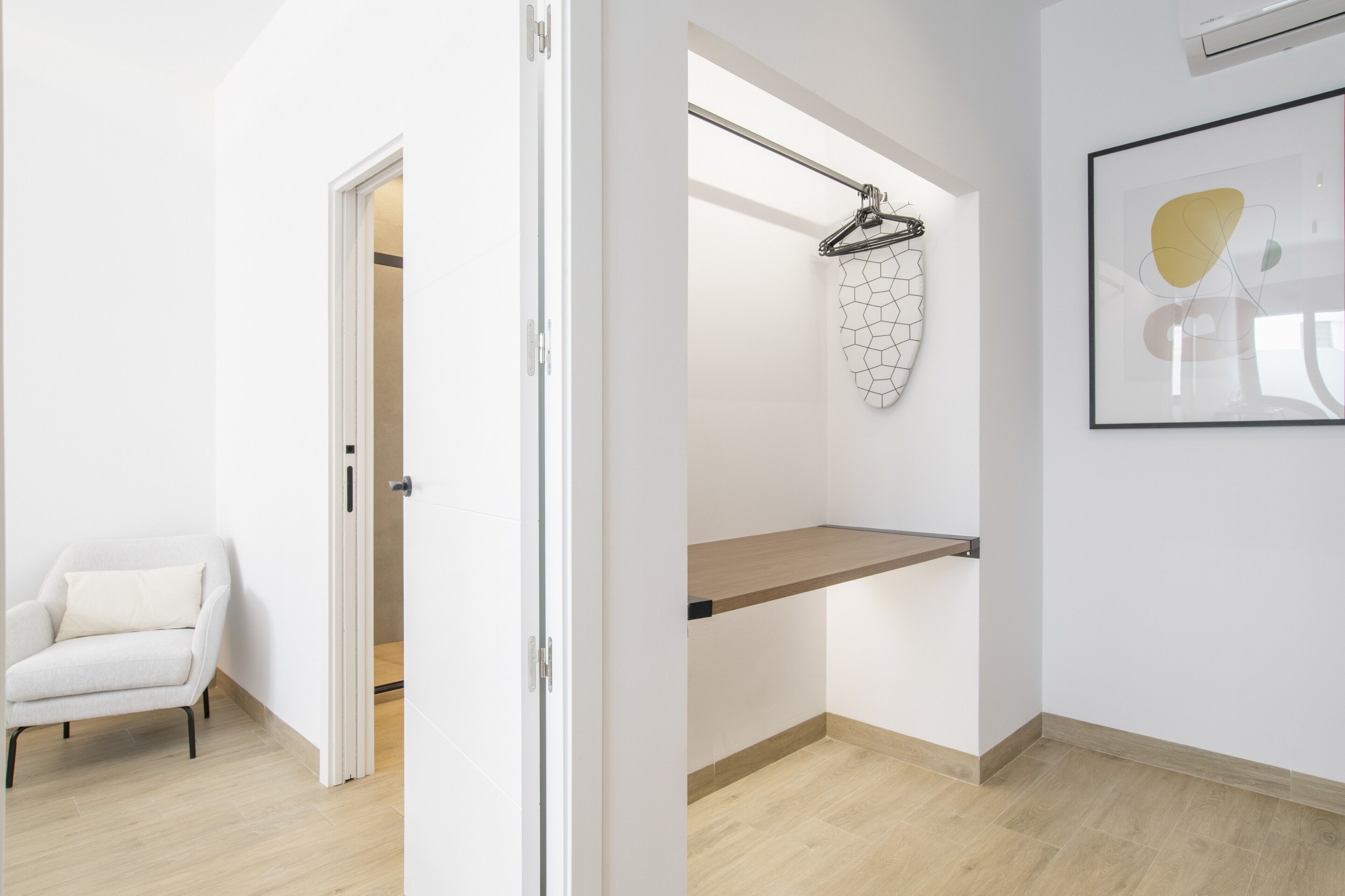 A modern room with a white chair, a coat rack with a geometric-patterned item, a wooden shelf, and a framed abstract artwork on the wall.