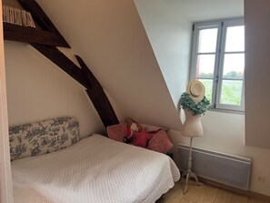 A bedroom with a sloped ceiling, a bed with a quilted cover, a radiator, and a window with a view of greenery.