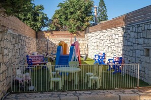Ein umzäunter Spielbereich im Freien mit Rutsche und Klettergerüst, umgeben von Steinmauern und einem Rasen.