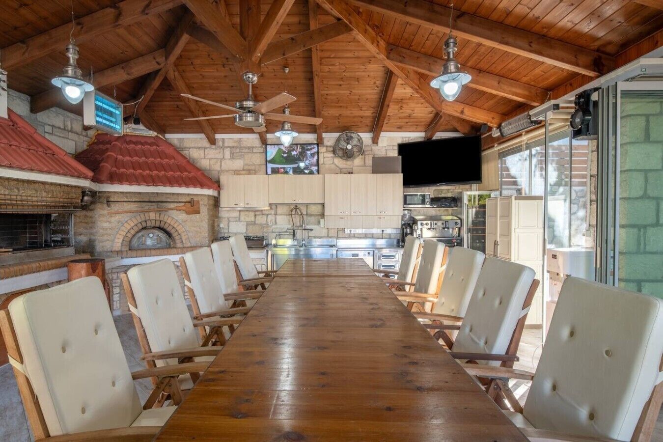 Une longue table à manger en bois, avec des chaises, une cheminée en pierre, un coin cuisine et un ventilateur de plafond.