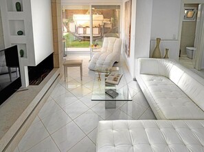 A modern living room with a white sofa, a glass coffee table, and a white armchair. There is a glass door leading to an outdoor area with a table and chairs. - Serenity Bliss Villa (Gouves)