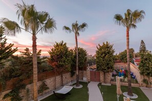 A landscaped outdoor area with palm trees, a brick wall, and a pathway leading to a seating area. - Serenity Bliss Villa (Gouves)