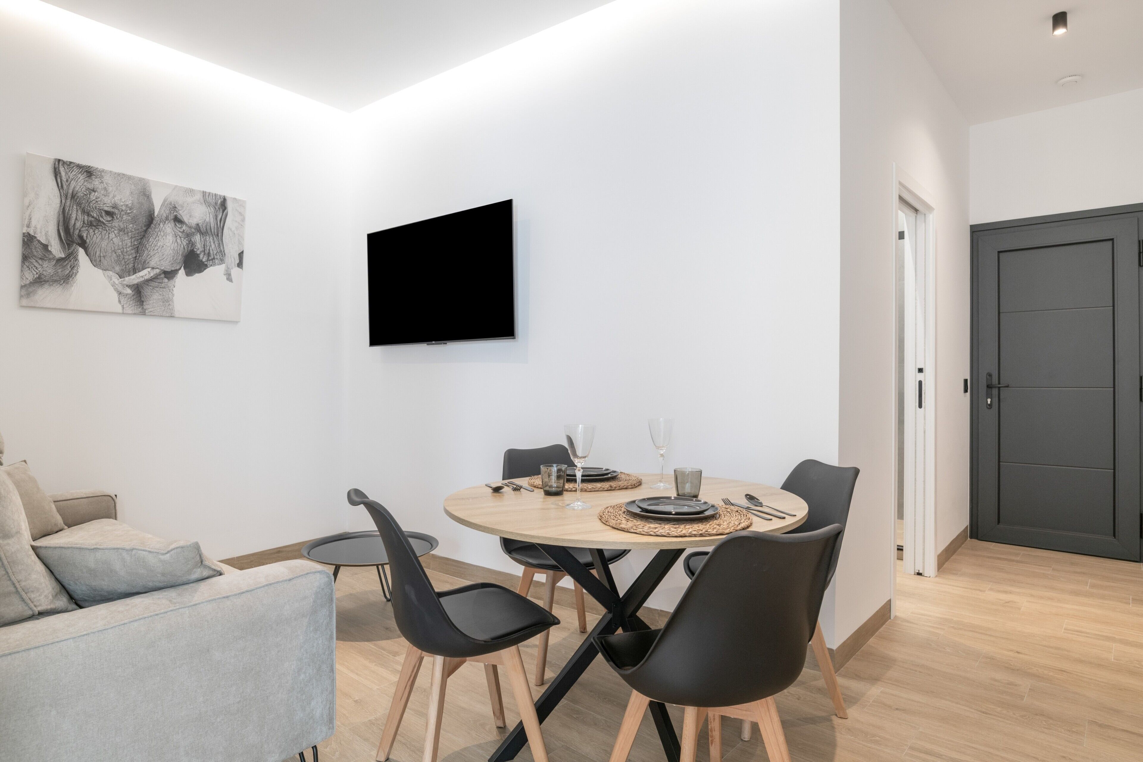 A modern living room with a round dining table, black chairs, a grey sofa, a flat-screen TV, and a large wall art piece.