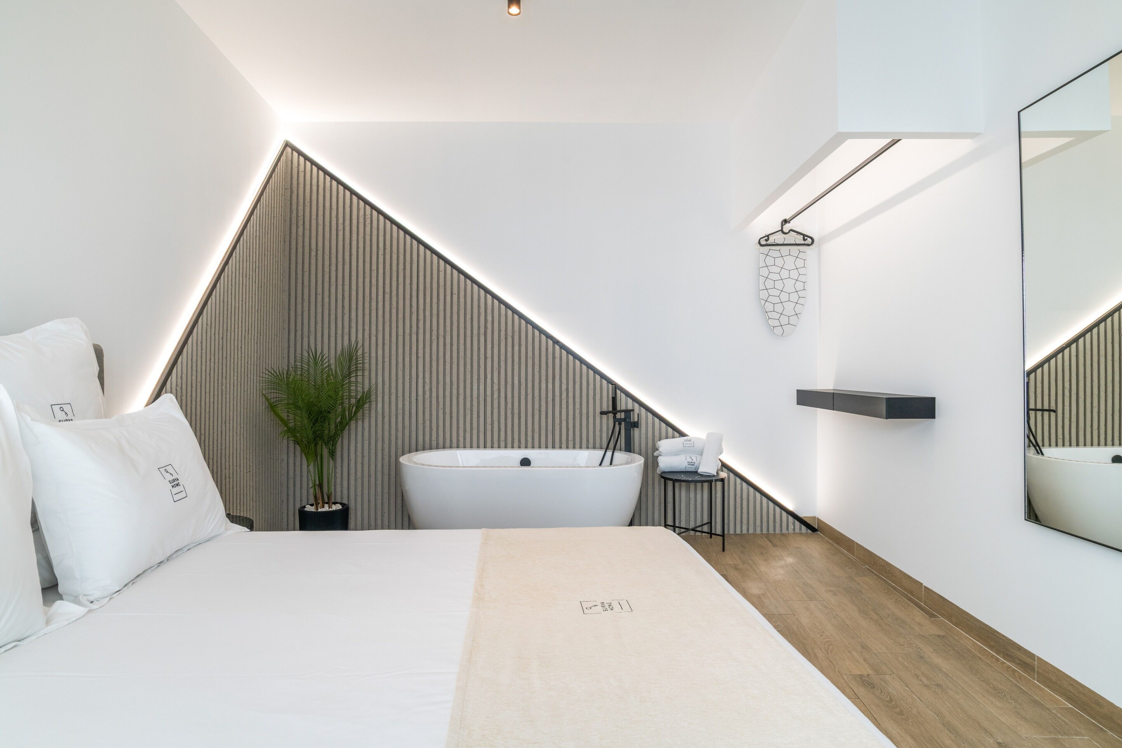 Un dormitorio moderno con una cama grande, una bañera independiente, una planta en maceta, un espejo y suelo de madera.