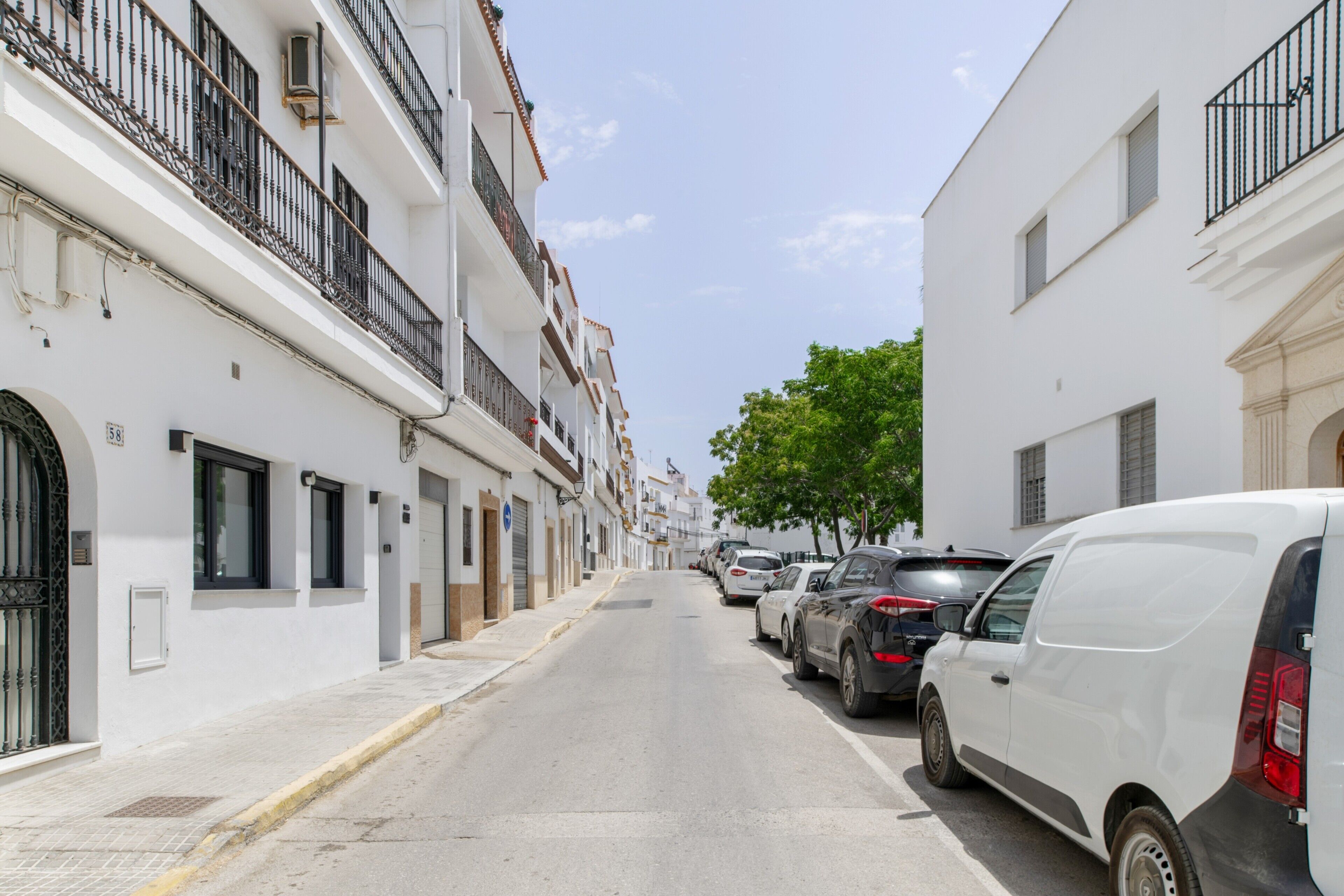 A narrow street lined with white buildings, parked cars, and a tree.