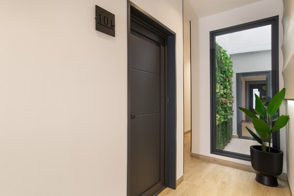 Un couloir avec une porte sombre, une plaque indiquant le numéro, une fenêtre donnant sur un espace verdoyant et un pot de fleurs.