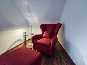 Un sillón rojo y un reposapiés en una esquina de una habitación con piso de madera y paredes blancas.