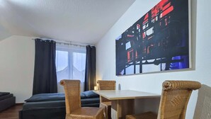 A room with a dining table, four wicker chairs, a sofa, and a large abstract painting on the wall. - Alpha Apartments: Grand Space | 6 Beds & Parking (Datteln)