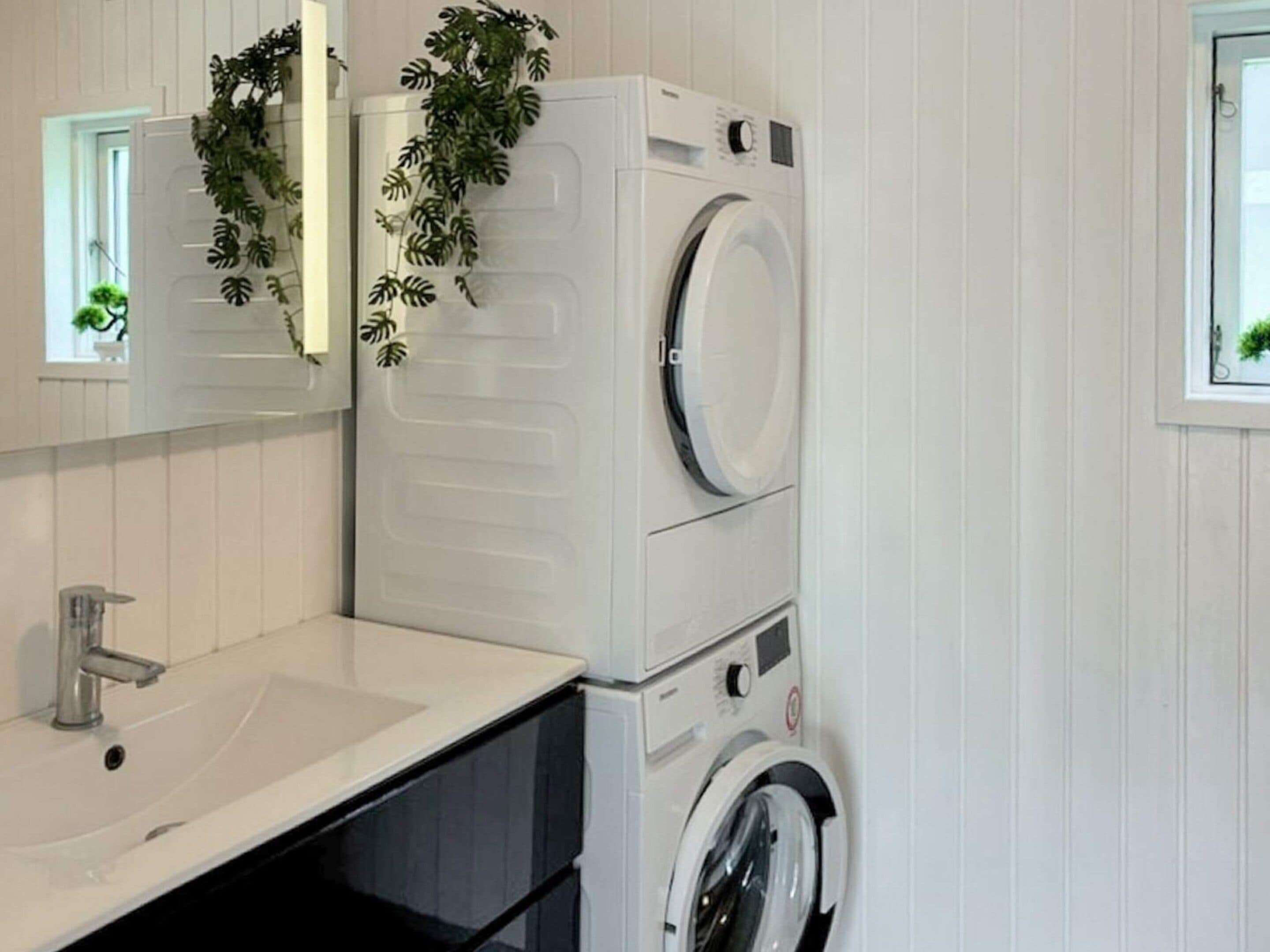 Un espace buanderie compact comprenant une machine à laver et un sèche-linge superposés, un évier et un miroir agrémenté d’une plante.