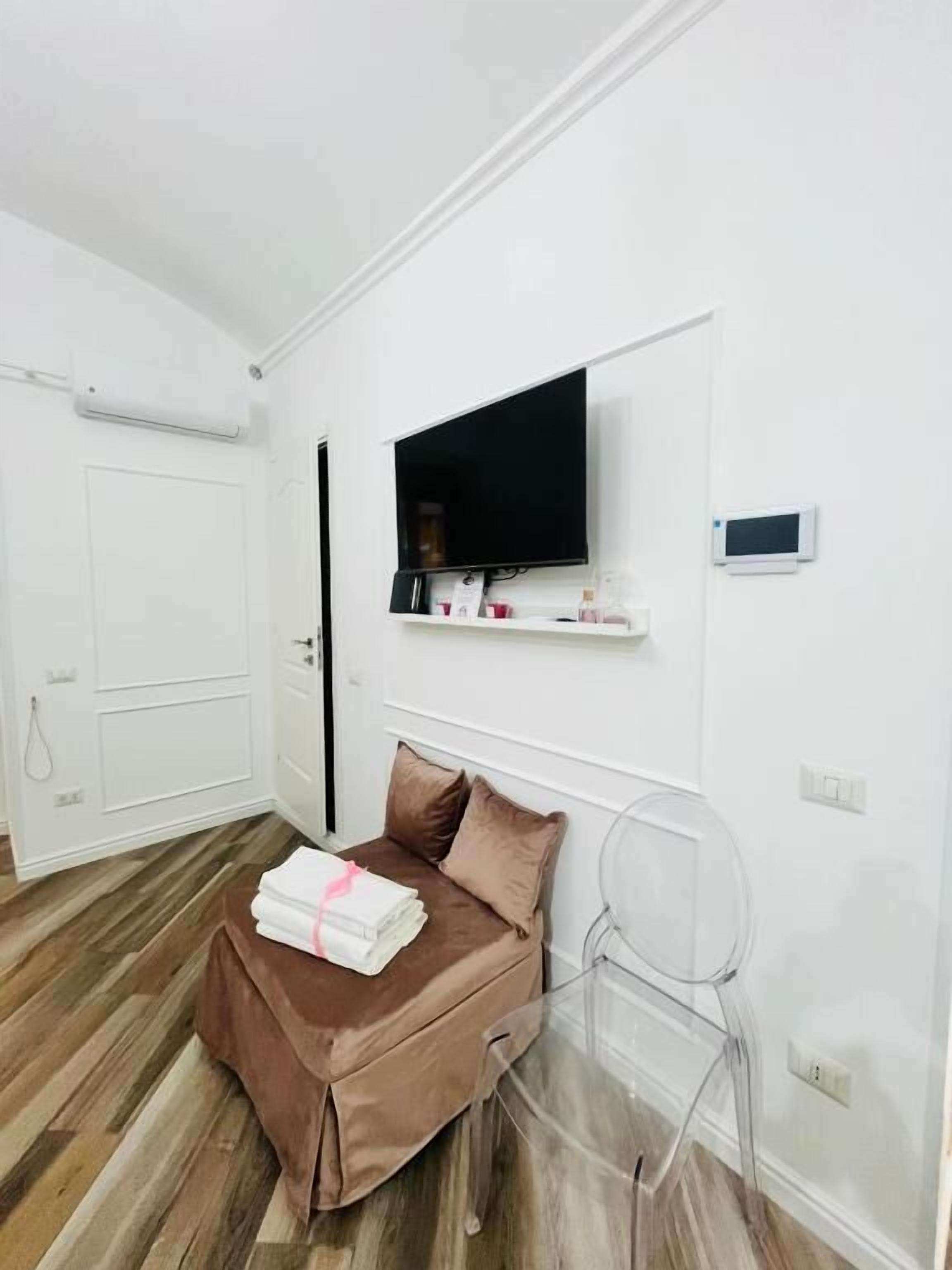A hotel room with a bed, a TV mounted on the wall, a small shelf with toiletries, and a clear chair.
