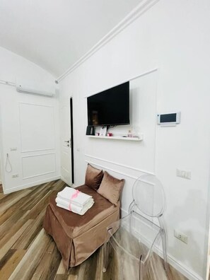 A hotel room with a bed, a TV mounted on the wall, a small shelf with toiletries, and a clear chair.