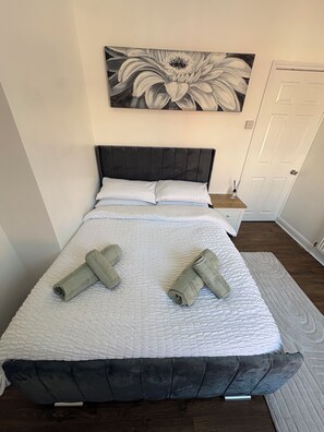 A neatly made bed with a white quilt and dark headboard, two rolled towels, a nightstand with a lamp, and a floral painting above the bed. - Spacious 4 Bedroom Home With Free Parking (Preston)