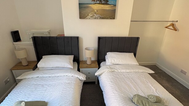 Ein Doppelzimmer mit zwei Betten, einem Nachttisch, einer Lampe, einem Gemälde und einem hölzernen Kleiderhaken an der Wand.