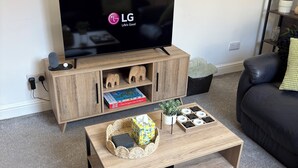 Ein Wohnzimmer mit Flachbildfernseher auf einem Holzboden, einem Couchtisch mit dekorativen Gegenständen, einem Bücherregal und einem schwarzen Ledersofa.