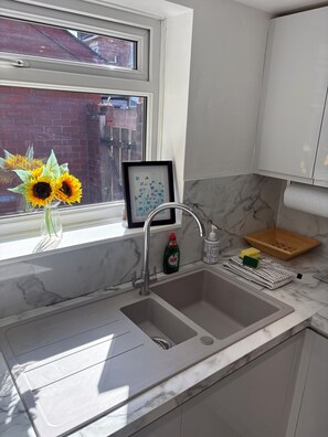 A kitchen with a double sink, a window with a view of a brick building, a framed picture, and a bottle of Fairy dish soap. - Spacious 4 Bedroom Home With Free Parking (Preston)