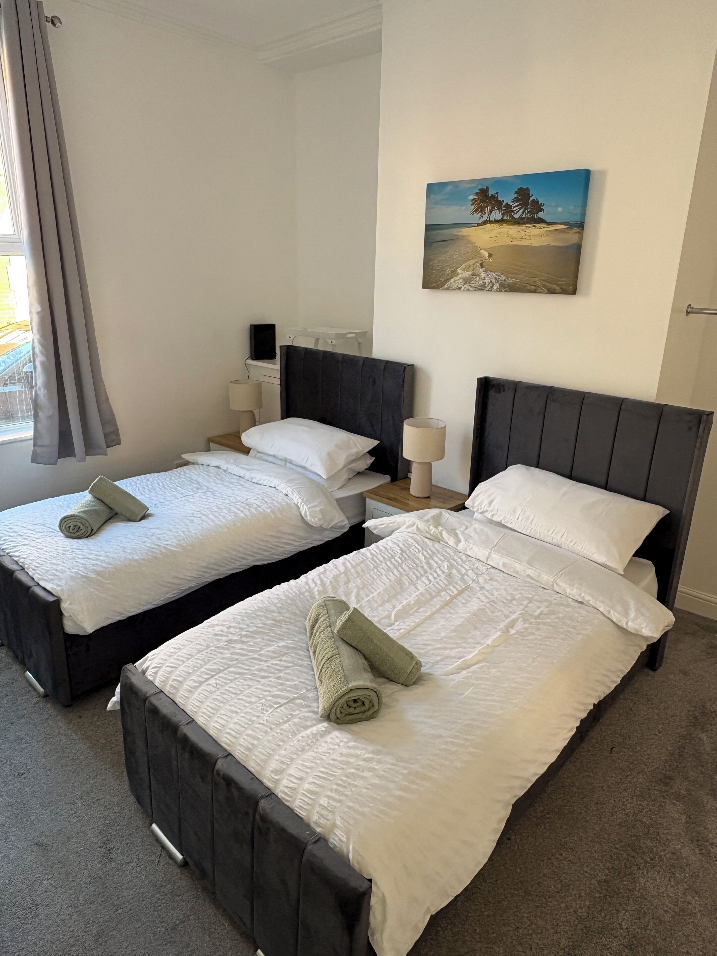 A hotel room with two beds, each with a dark headboard, white bedding, and a rolled towel. There is a painting of a beach scene on the wall.