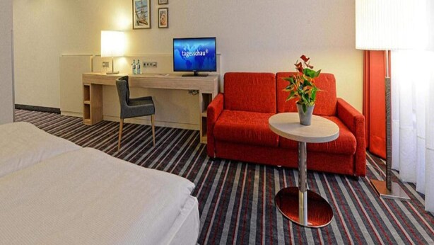 Et hotelværelse med en seng, et skrivebord med en computer, en rød sofa, et lille bord med en plante og en lampe.