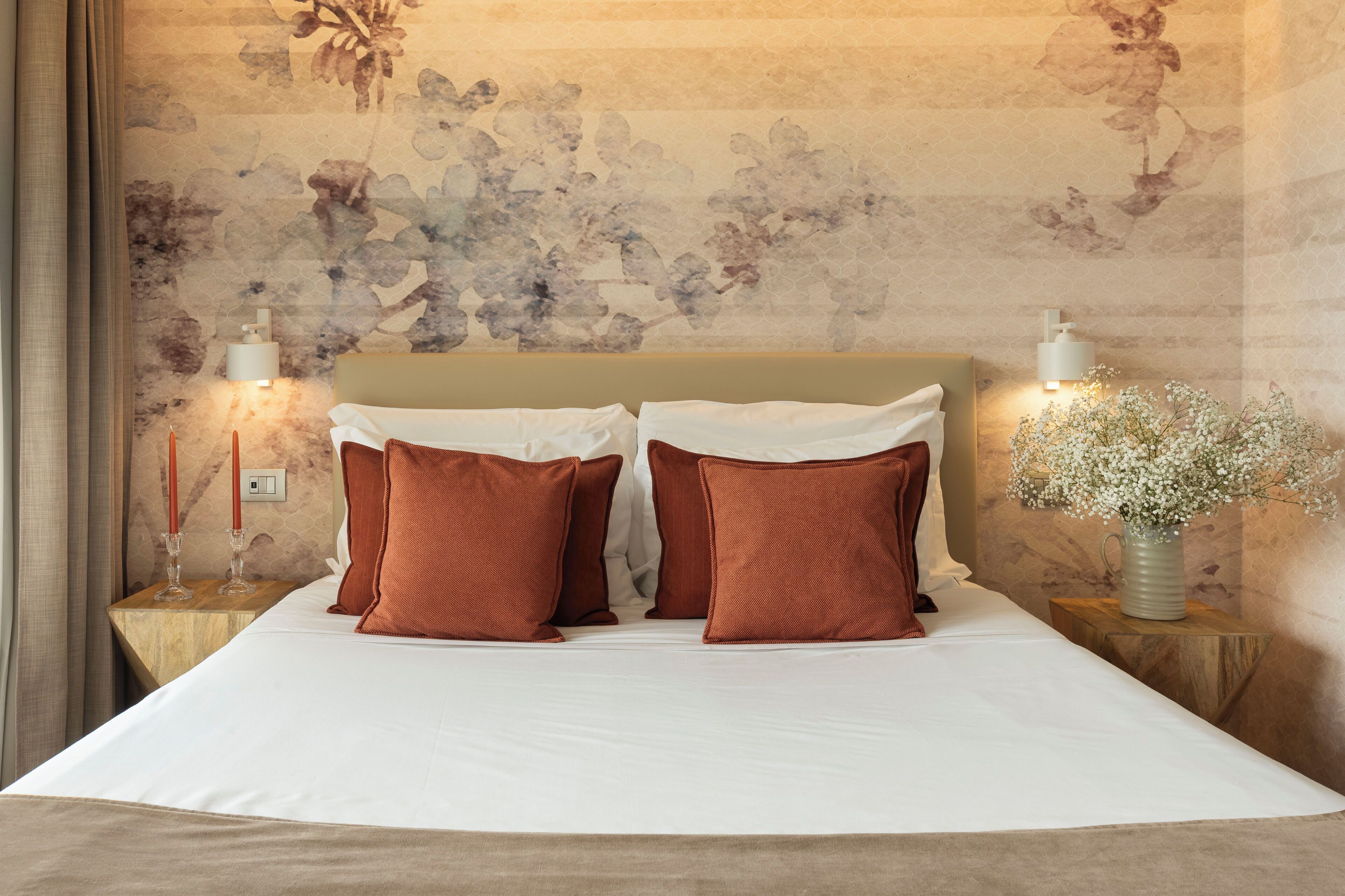 A neatly made bed with white linens and two brown pillows, a vase with white flowers on a nightstand, and a wall with a floral pattern.