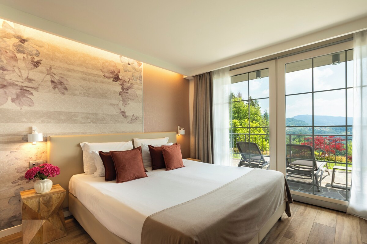 Ein Hotelzimmer mit einem großen Bett, einem Nachttisch mit Blumen in einer Vase, einer Wandleuchte und einem Balkon mit Stühlen und Blick ins Grüne.