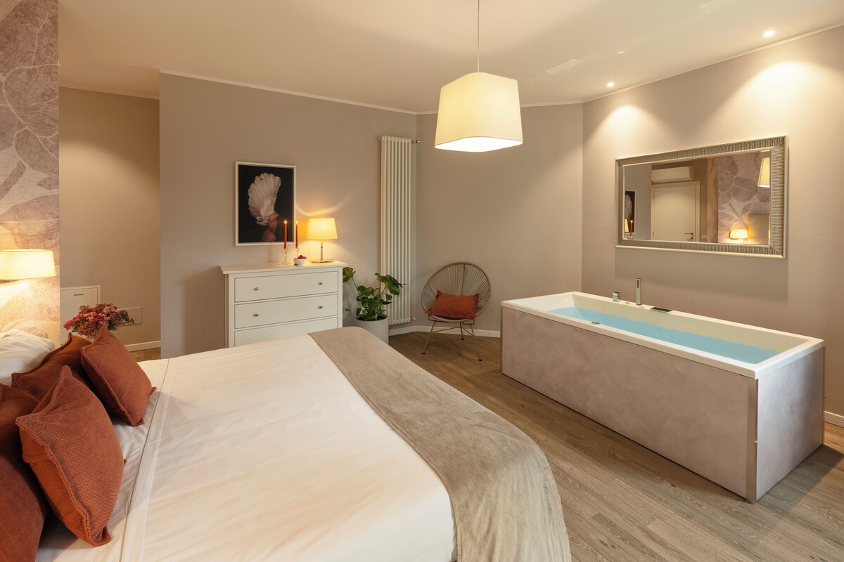 Ein modernes Schlafzimmer mit einem großen Bett, einem Nachttisch mit Lampe, einer Kommode, einem Sessel und einer großen Spiegel sowie einer Badewanne.