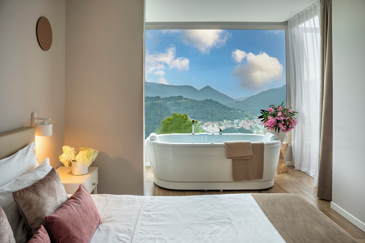 Ein Schlafzimmer mit einem großen Bett, einer weißen Badewanne, einem Nachttisch mit Lampe und Blumen sowie Bergblick durch ein Fenster.