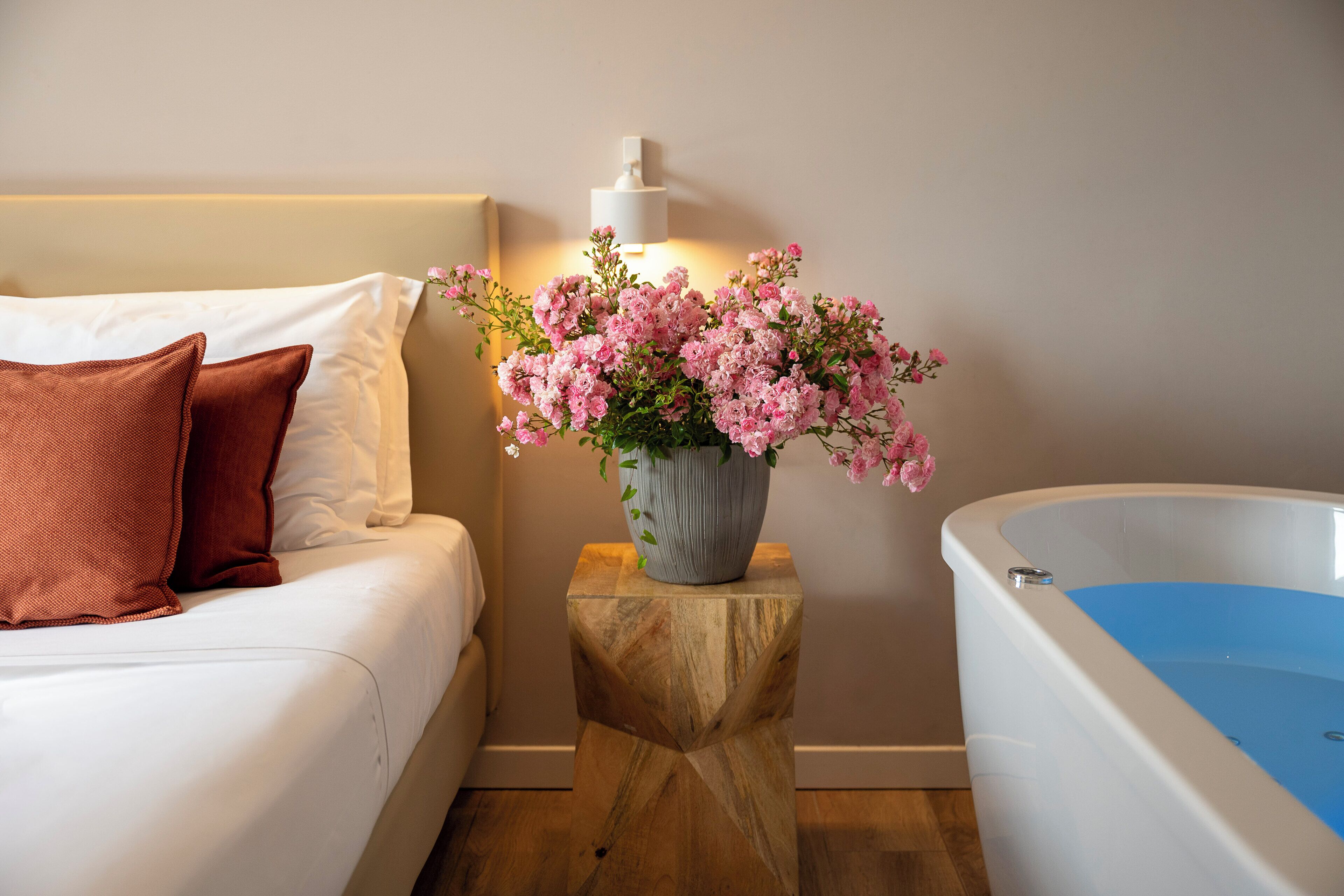A bedroom with a bed, a wooden nightstand with a vase of flowers, and a bathtub.