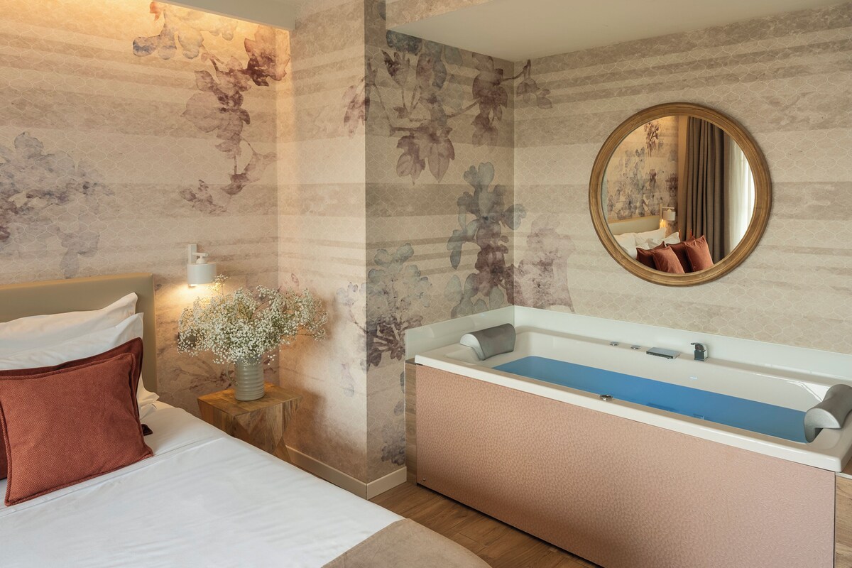 Ein Schlafzimmer mit einem Bett, einem Nachttisch mit einer Vase, einem großen runden Spiegel und einer Badewanne.