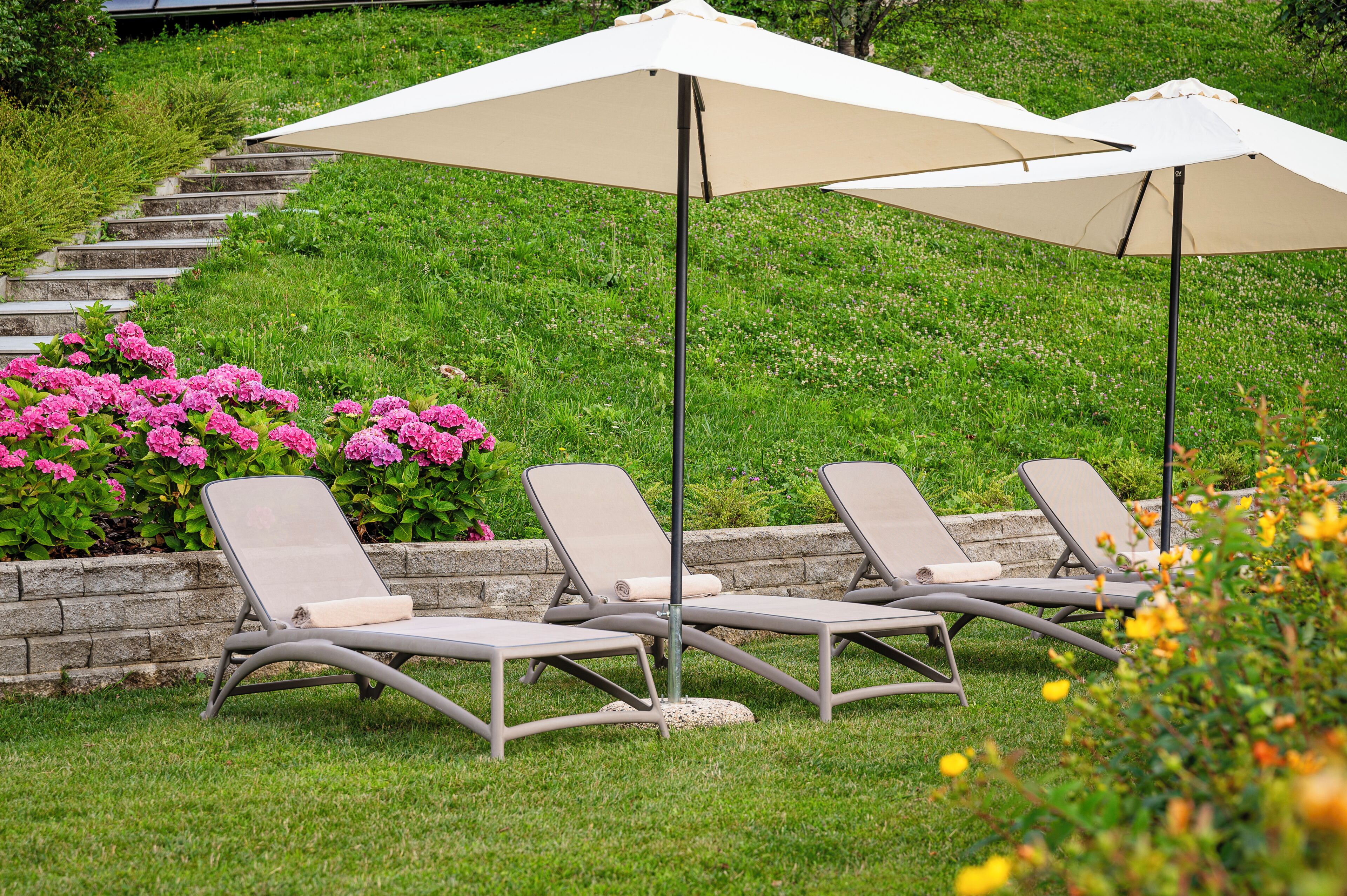 Outdoor lounge chairs with umbrellas, a stone retaining wall, and pink flowers.