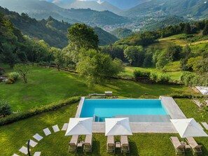 Ein Pool mit Liegestühlen und Sonnenschirmen, umgeben von üppiger grüner Landschaft und Bergen im Hintergrund.