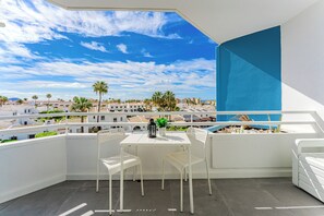A balcony with a white table and chairs, a potted plant, and a view of a coastal town with palm trees and buildings. - Home Holidays 423 (CN)