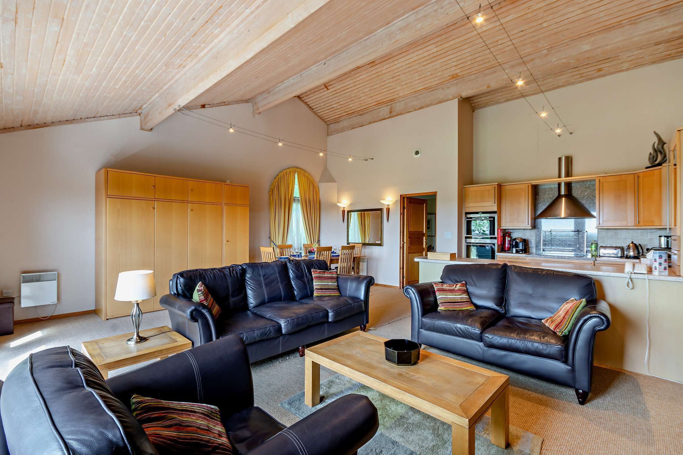 A spacious living area with leather sofas, a wooden coffee table, and a kitchen in the background.