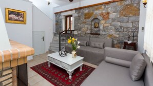 A living room with a stone wall, a sofa, a coffee table with flowers, a staircase, and a chandelier. - Pleades Seaside Asterope Gem in Mani Near the Sea (West Mani)