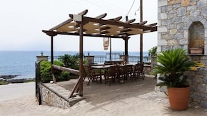 An outdoor seating area with tables and chairs, a pergola, a stone building with a wooden door, and a view of the sea. - Pleades Seaside Asterope Gem in Mani Near the Sea (West Mani)