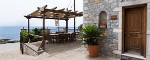 Ein Sitzbereich im Freien mit Tischen und Stühlen, einer Pergola, einem Steinhaus mit Holztür und Blick aufs Meer.