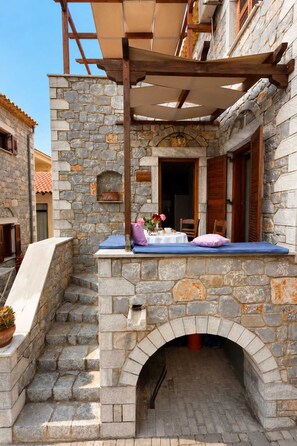 Ein Sitzbereich im Freien aus Stein mit integriertem Kamin, einer hölzernen Pergola darüber und Blick auf das angrenzende Gebäude.
