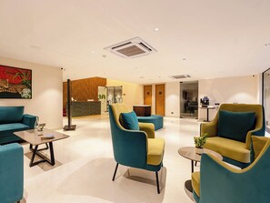 Um hall de hotel moderno com poltronas verde-água e mostarda, uma mesa de centro e um balcão de recepção.