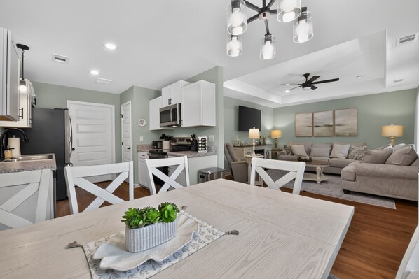 A modern kitchen with white cabinets, a dining area with a wooden table, and a living room with a sofa and a TV.