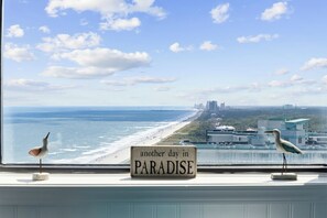 Blick auf einen Strand und eine Stadtansicht durch ein Fenster, mit einem Schild, auf dem 'another day in PARADISE' steht.