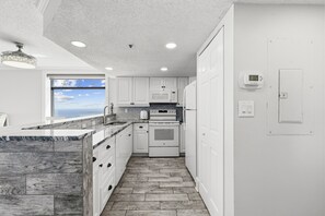 A modern kitchen with white cabinetry, a marble countertop, a built-in oven, and a view of the ocean through a window. - Beach Colony Oceanfront 3BR Corner Retreat (Myrtle Beach)